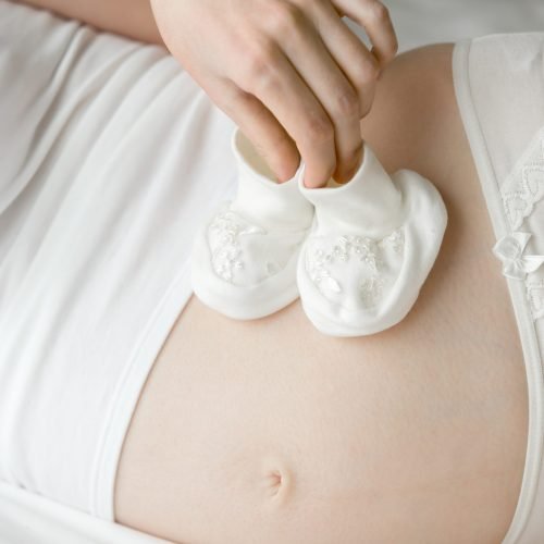 Mom and her future child. Young pregnant model lying on the bed and holding cute white baby booties on her belly. Close-up. Maternity concept